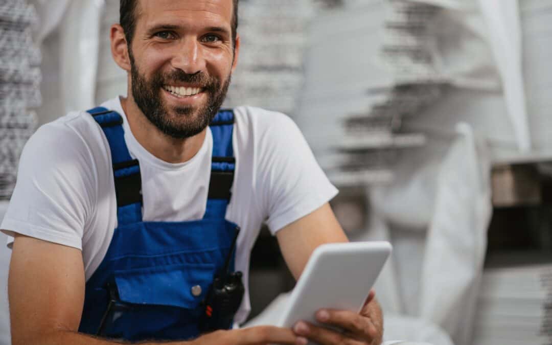 A smiling worker in a blue overall holding a tablet, documenting production steps with a digital manufacturing traveler in a modern factory setting.