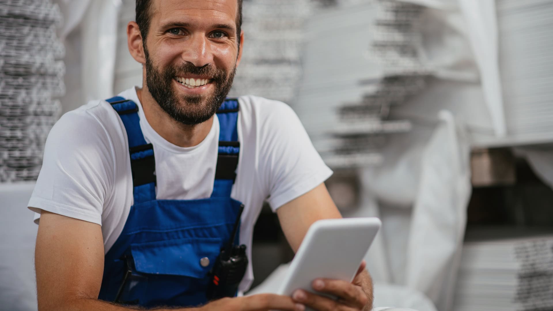 A smiling worker in a blue overall holding a tablet, documenting production steps with a digital manufacturing traveler in a modern factory setting.